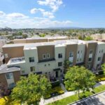 Aerial view of a row of modern, multi-story townhouses with beige and gray facades, green front lawns, and trees along a residential street.
