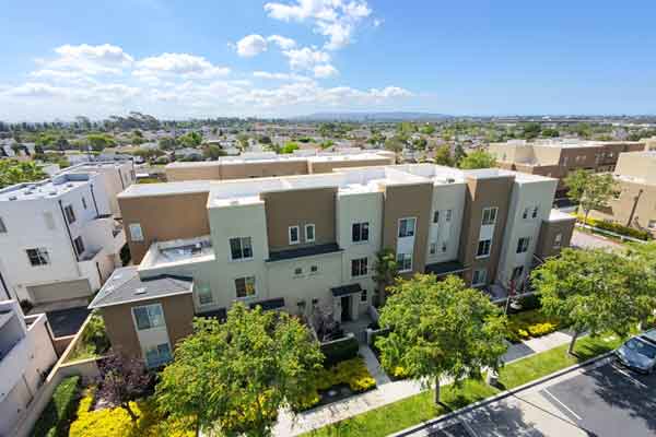 Aerial view of a row of modern, multi-story townhouses with beige and gray facades, green front lawns, and trees along a residential street.