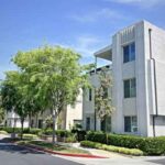 Modern beige apartment complex with trimmed hedges and trees along a sunny street.
