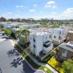 Terrace-and-Courts-drone - Three Sixty At South Bay Aerial view of a modern townhouse complex with white buildings, palm trees, and a tree-lined street under a blue sky.