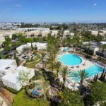 Aerial view of a sunny resort with a large, irregular pool surrounded by palm trees and white buildings.