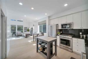 Bright open-plan kitchen and living room with white cabinets, stainless steel appliances, and a central island with shelves.