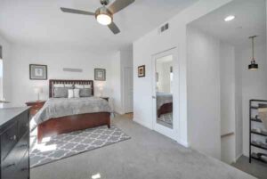 Spacious white-themed bedroom with a wooden bed, two nightstands, and a geometric rug under the foot of the bed; ceiling fan overhead.