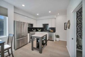 Bright modern kitchen with white cabinets and stainless steel appliances, a dark wood island, light flooring, and a doorway to a tiled bathroom.