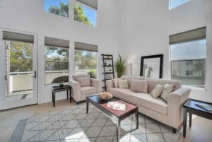 Bright, airy living room with beige sofas, a glass coffee table, and a geometric rug beneath a sunlit seating area. Large windows and a glass door bring in natural light.