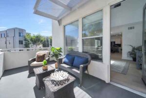 Sunny balcony with wicker seating (two chairs with blue cushions) and a small coffee table with plants; large sliding doors reveal a living room indoors.